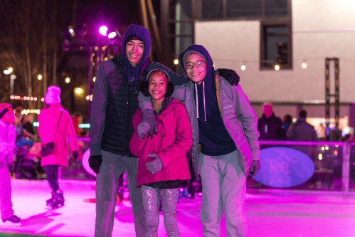 Three children smile together on an ice rink, surrounded by colorful lights and festive decorations.