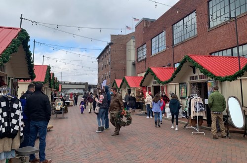 A bustling outdoor market with festive stalls, shoppers browsing, and holiday decorations along a brick pathway.
