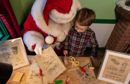 Santa Claus shows a young boy toy blueprints at a festive workshop, surrounded by wooden toys and plans.