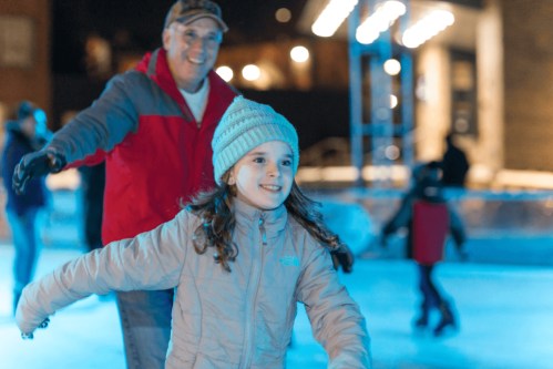 A man and a girl skate joyfully on an ice rink, surrounded by other skaters in a festive atmosphere.