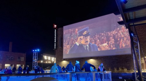 A large screen displays a scene from a movie while people skate on an ice rink at night, surrounded by colorful lights.