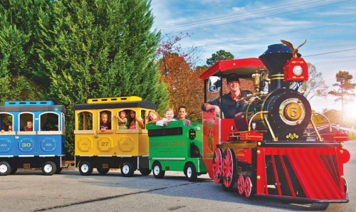 A colorful miniature train with children riding, driven by an adult, set against a backdrop of trees and a clear sky.