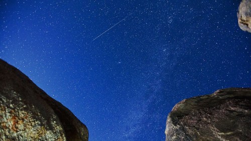 A starry night sky with a shooting star, framed by large rocks in the foreground.