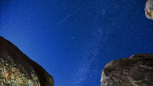 A starry night sky with a shooting star, framed by large rocks on either side.