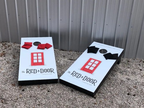 Two cornhole boards with red and black bags, featuring a design of a red door, set on a gravel surface.