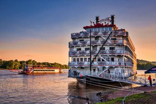 A large riverboat docked by the shore at sunset, with another boat passing in the background.