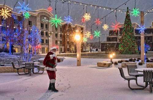 Santa Claus stands in a snowy plaza decorated with colorful lights and a Christmas tree, surrounded by festive decorations.