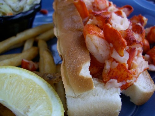 A lobster roll filled with chunks of lobster meat, served with fries and a lemon wedge on a blue plate.