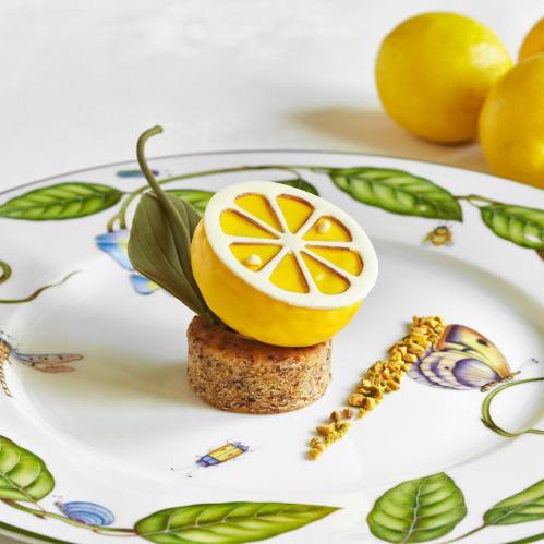 A decorative dessert featuring a lemon slice atop a round cake, garnished with a leaf and crumbs, on a patterned plate.