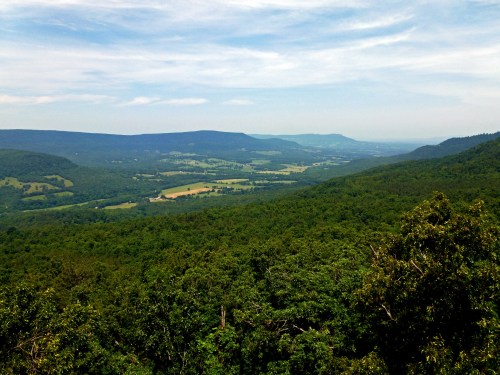 A panoramic view of lush green valleys and distant mountains under a blue sky with scattered clouds.