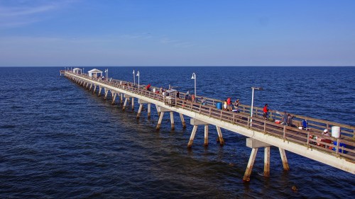 A long wooden pier extends over calm blue waters, with people fishing and enjoying the view under a clear sky.