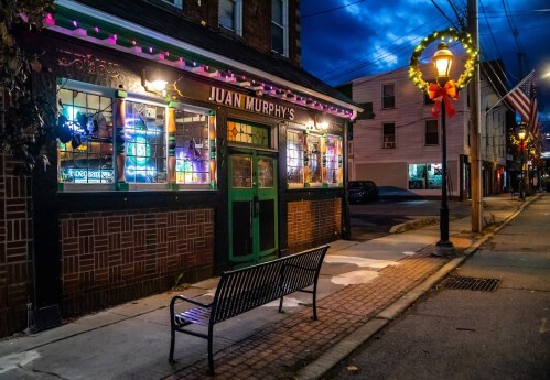 A cozy pub with colorful neon signs, festive lights, and a bench outside, set against a twilight sky.