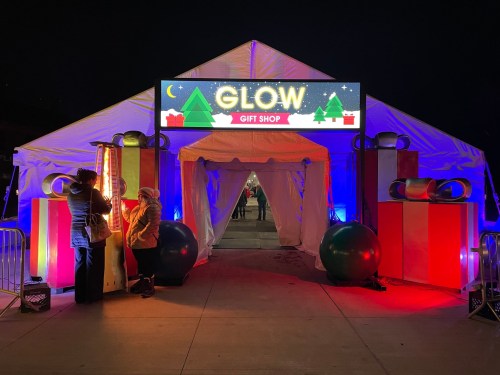 A festive tent entrance with a "GLOW GIFT SHOP" sign, decorated with large gift boxes and colorful lights.