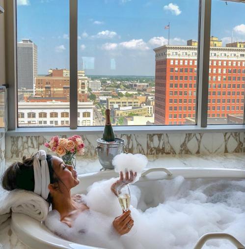 A person relaxes in a bubble bath with a glass of champagne, enjoying a city view through a large window.