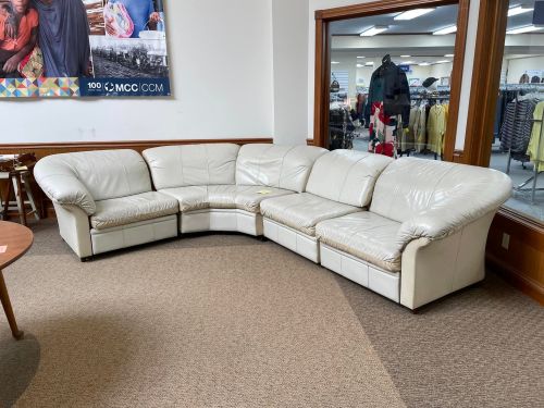 A white leather sectional sofa arranged in a corner of a store, with a textured carpet and clothing racks in the background.