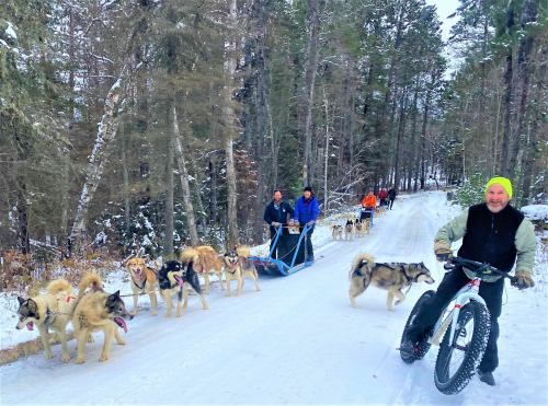 A group of people sledding with dogs on a snowy trail, surrounded by trees in a winter landscape.