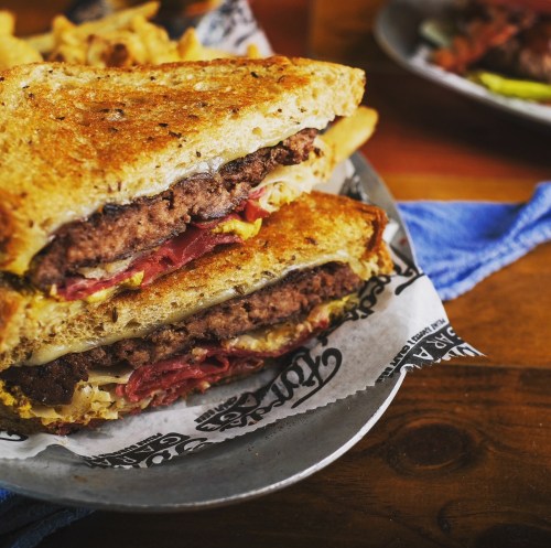 A close-up of a stacked sandwich with beef patty, cheese, and toppings, served with fries on the side.