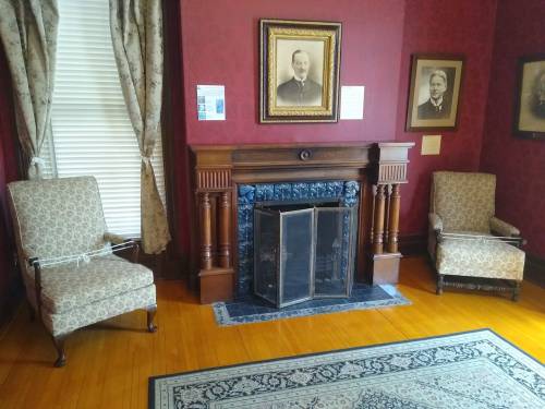 A cozy room with two patterned chairs, a fireplace, and framed portraits on red walls. A decorative rug is on the floor.