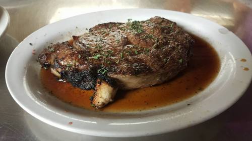 A juicy, grilled ribeye steak on a white plate, garnished with herbs and resting in a savory sauce.
