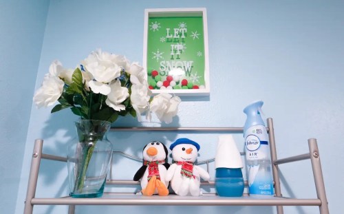 A shelf with a green "Let It Snow" sign, flowers, and two penguin plush toys wearing scarves.