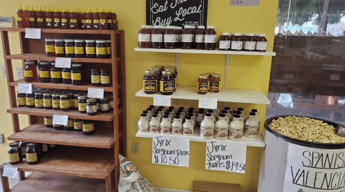 Shelves displaying various jars of honey, syrup, and preserves with price tags in a local market setting.