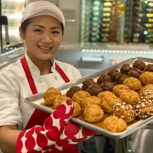 A smiling chef in a white uniform and red apron holds a tray of freshly baked pastries.