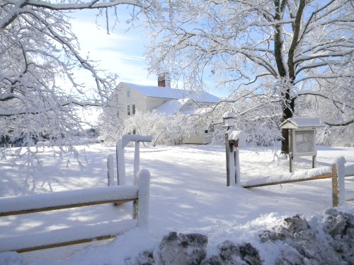 A snow-covered landscape featuring a white house surrounded by trees and a wooden fence in a winter setting.