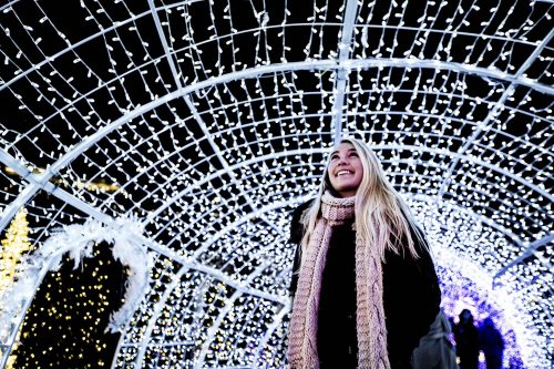 A smiling woman stands under a canopy of twinkling holiday lights, creating a festive atmosphere.