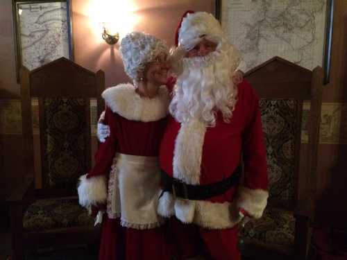 Santa Claus and Mrs. Claus pose together, both dressed in traditional holiday attire, smiling in a cozy setting.