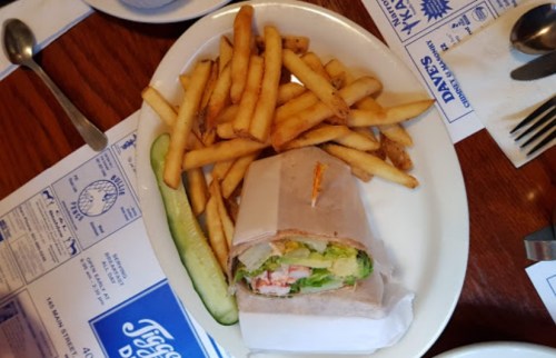 A plate with a wrapped sandwich, fries, and a pickle on the side, set on a wooden table with menus underneath.