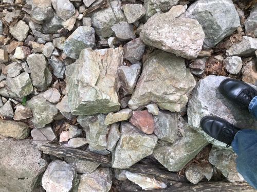A person's feet in black shoes standing on a rocky, uneven surface with various sizes of stones and pebbles.
