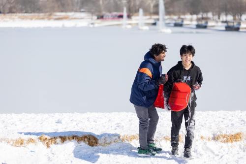 Two boys play in the snow near a frozen lake, one holding a sled and the other laughing as they interact.