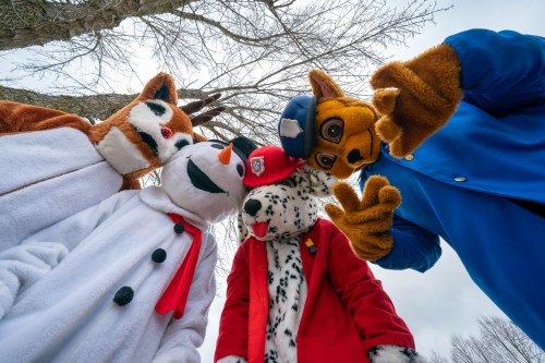 Four costumed characters, including a snowman and animals, pose playfully under bare trees against a cloudy sky.