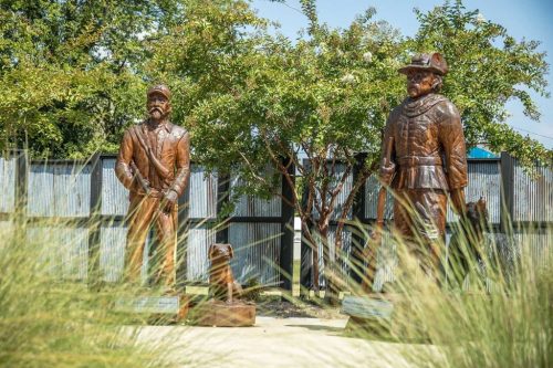 Two bronze statues of historical figures stand in a landscaped area, surrounded by greenery and tall grass.