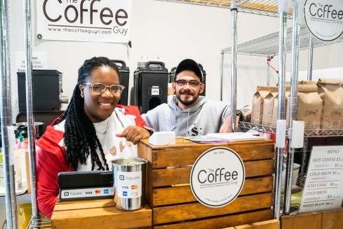 Two baristas smile behind a wooden counter at a coffee stand, with bags of coffee and a menu in the background.