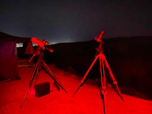 Two telescopes set up on tripods in a dark, red-lit outdoor setting, with a distant horizon and stars visible.