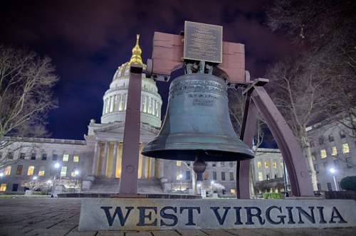 A large bell in front of the West Virginia State Capitol building at night, illuminated by surrounding lights.