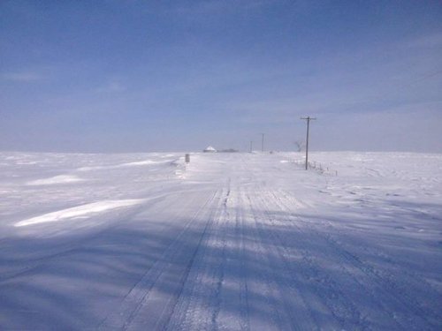A snowy landscape with a road disappearing into the distance, flanked by snow-covered fields and a clear blue sky.