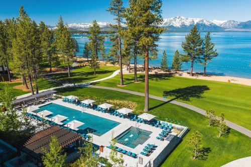 Aerial view of a resort pool surrounded by green lawns, tall trees, and a scenic lake with mountains in the background.