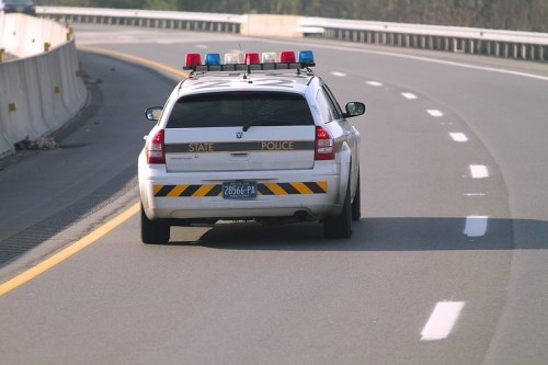 A state police car with flashing lights drives on a highway, viewed from the rear.