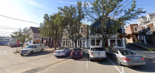 A street view of a small town with parked cars and shops, surrounded by trees under a clear blue sky.
