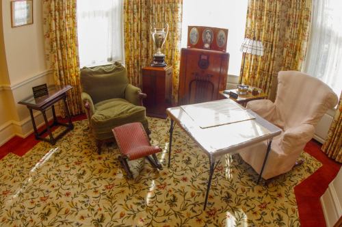 A cozy vintage room with patterned curtains, a trophy, an old radio, and two armchairs on a floral carpet.