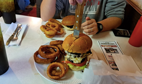 A large burger with toppings and a knife in it, accompanied by onion rings on a plate at a diner table.