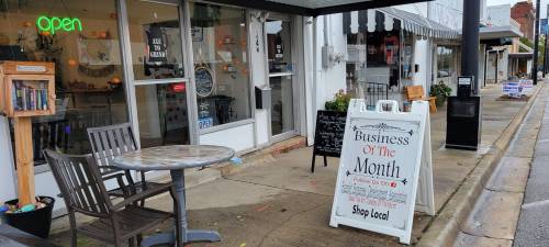 A storefront with an "Open" sign, outdoor seating, and a "Business of the Month" sign promoting local shopping.