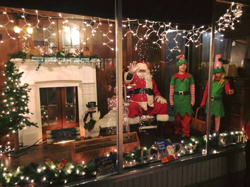 A festive window display featuring Santa Claus and two elves surrounded by Christmas lights and decorations.