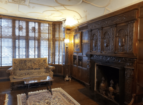 A cozy room with wooden paneling, a vintage sofa, a fireplace, and ornate ceiling details, illuminated by soft light.