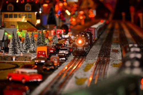 A miniature train set with a locomotive, cars, and festive decorations, surrounded by trees and buildings.