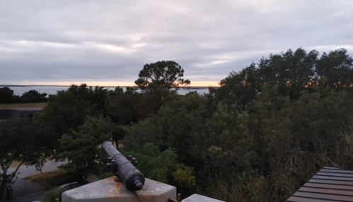 A view from a rooftop overlooking trees and a body of water at sunset, with a telescope in the foreground.