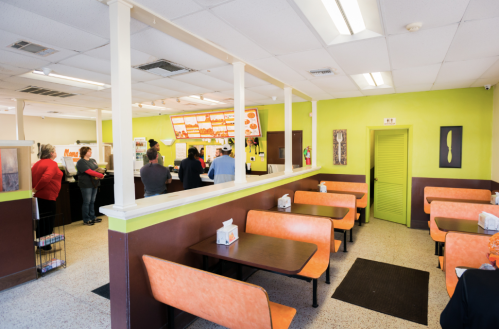 A colorful diner interior with orange booths, a counter, and customers waiting in line. Bright green walls add vibrancy.