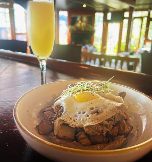 A bowl of savory dish topped with a fried egg, alongside a glass of mimosa in a bright, cozy restaurant setting.